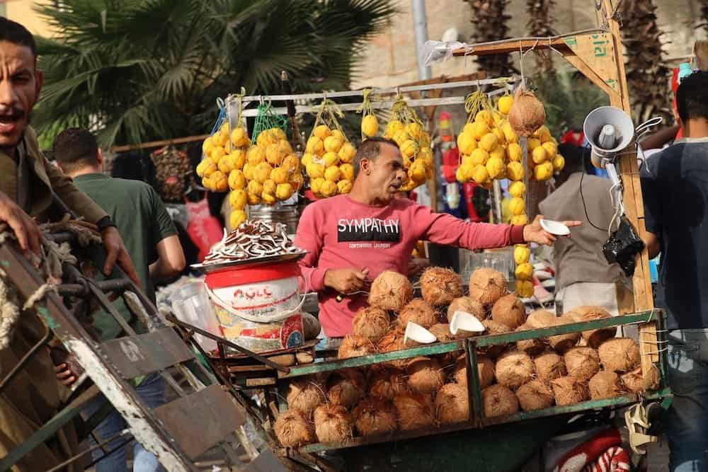 a man wearing a shirt that says "sympathy" is haggling over the price of coconuts he is vending with a row of lemons behind him. Learning how to haggle is a top Morocco travel tip. 