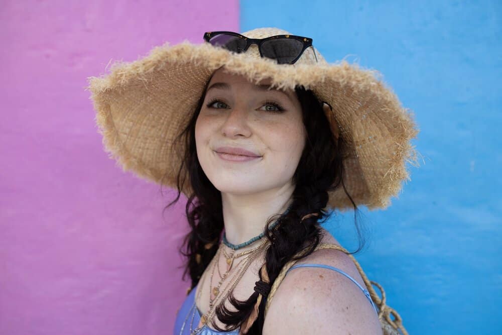 Blogger Katie Caf wearing a straw hat and two braids in her hair, photographed against a pink and blue wall at Malang's Rainbow Village. 