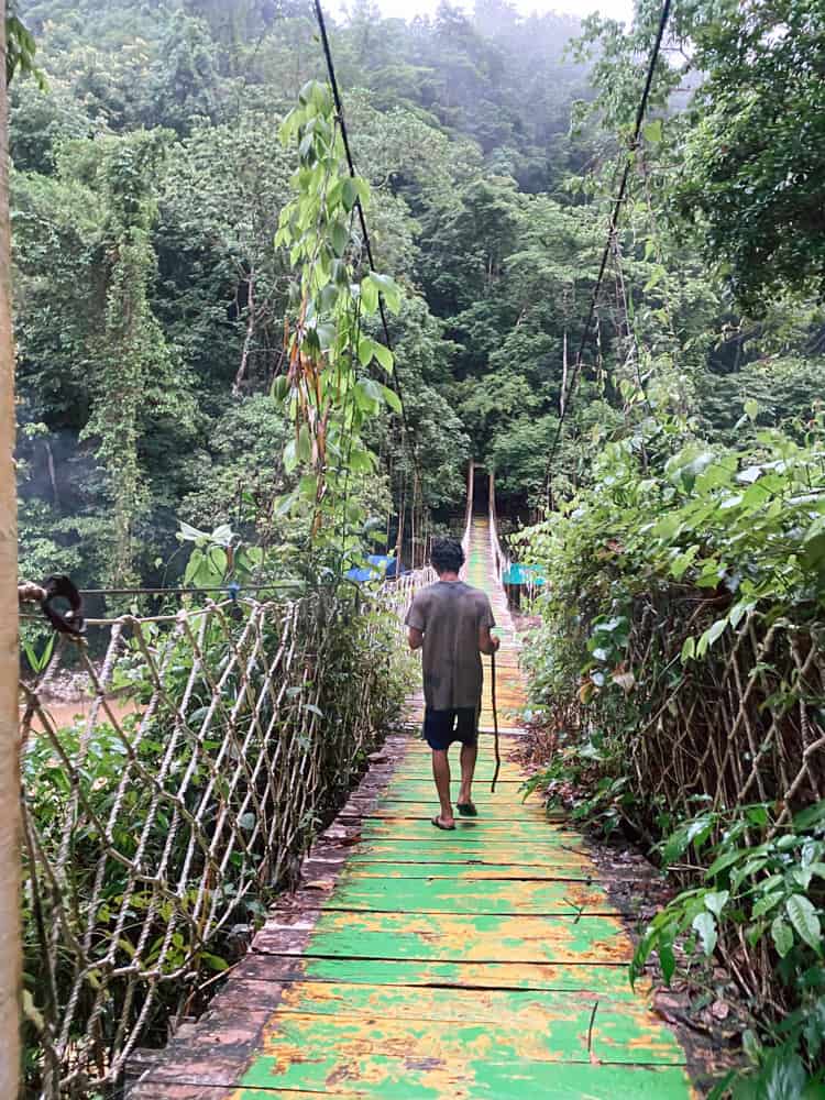 a tour guide at Cunca Wulang Waterfall in Labuan Bajo, Flores, crossing a bridge to reach the falls and carrying a walking stick.