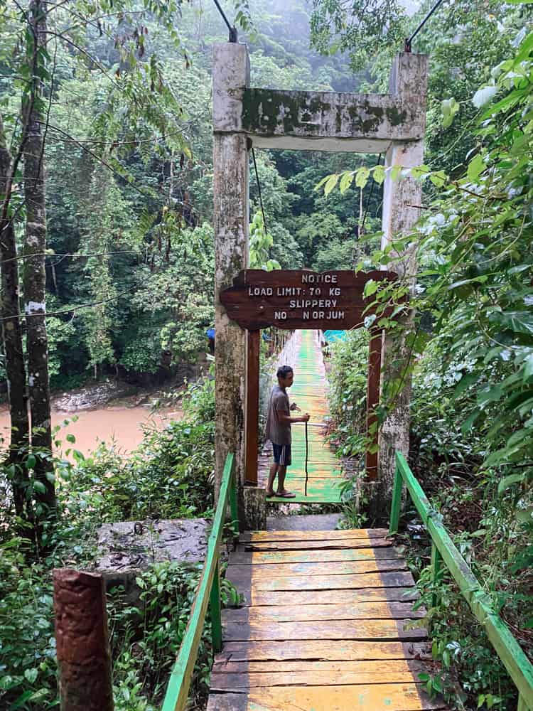 A tour guide standing with a walking stick at a run down bridge painted yellow. The bridge makes up the pathway to visit Cunca Wulang Waterfall.