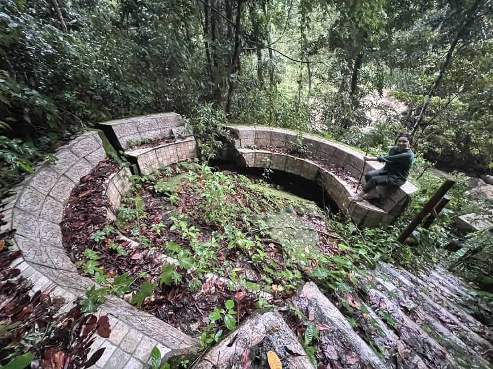 Blogger Katie Caf sitting on a broken stone pit that was once used as a seating area for tourist at Cunca Wulang Waterfall. The pit is overgrown with moss and weeds and is cracked in half.
