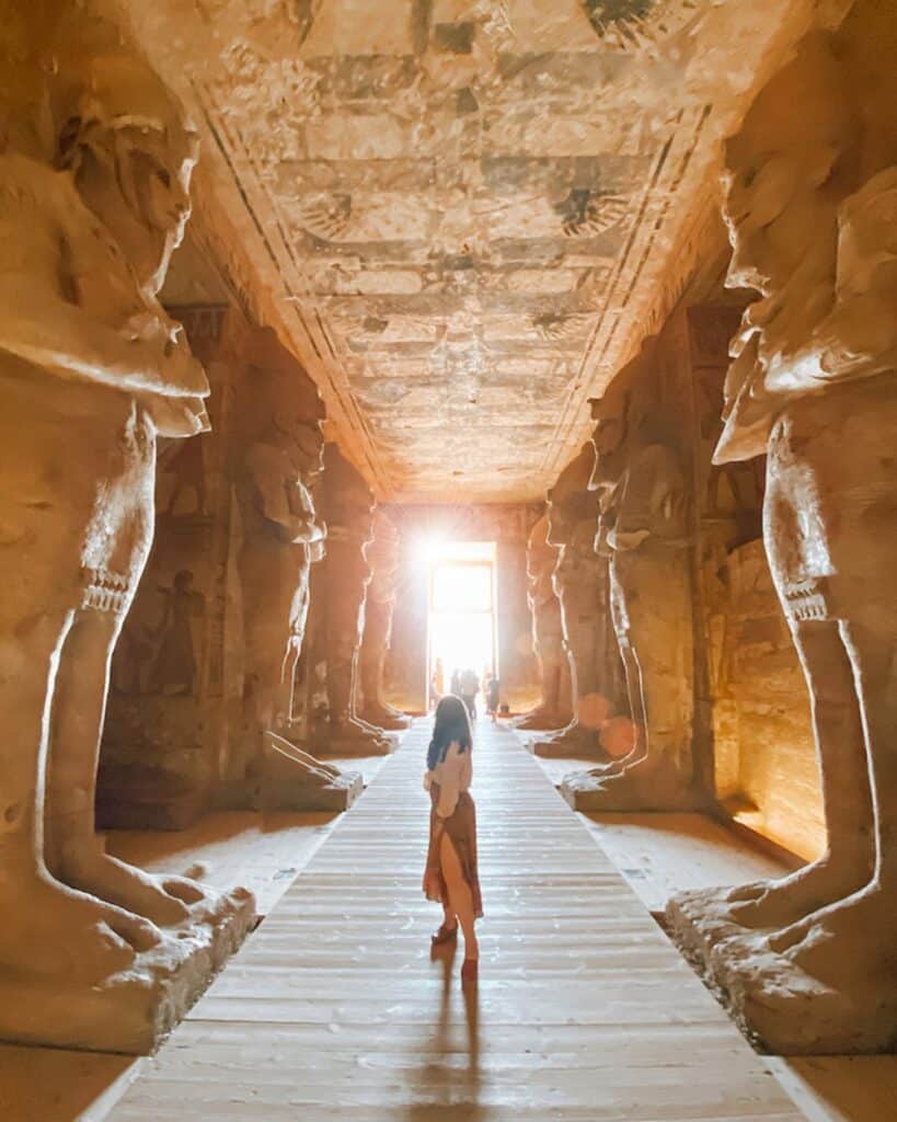 A girl staring at the entrance to Abu Simbel temple from the inside, a sun beam glaring at the door. overhead are large Pharonic sculptures 10x the size of a human acting as columns holding up the structure. Abu Simbel is one of many excursions people have on Nile River Cruises from Cairo to Luxor and Aswan.