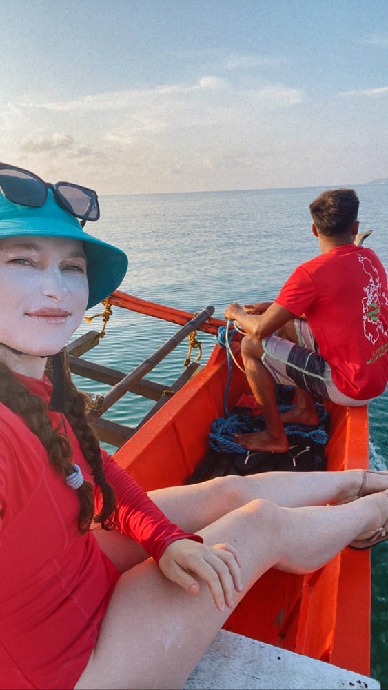 Blogger Katie Caf wearing white surfing sunscree, a red rashguard, blue bucket hat, and sunglasses on a boat. In the front of the boat, you can see the captain wearing a red shirt. 