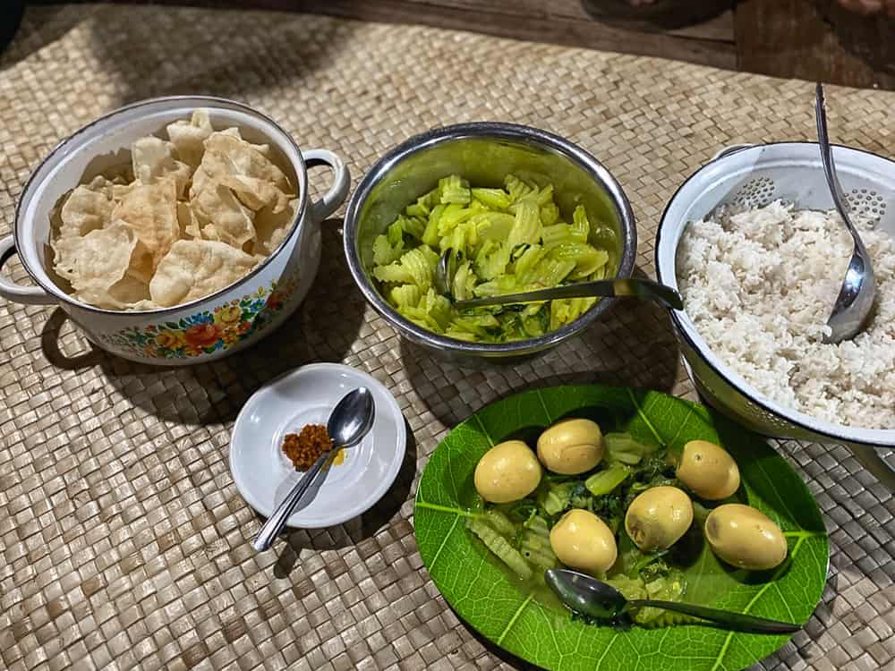 five bowls on a woven matt, one with eggs, one rice, one greens, and another chips. This is the family-style dinner guest are served at Wae Rebo Traditional village homestays.