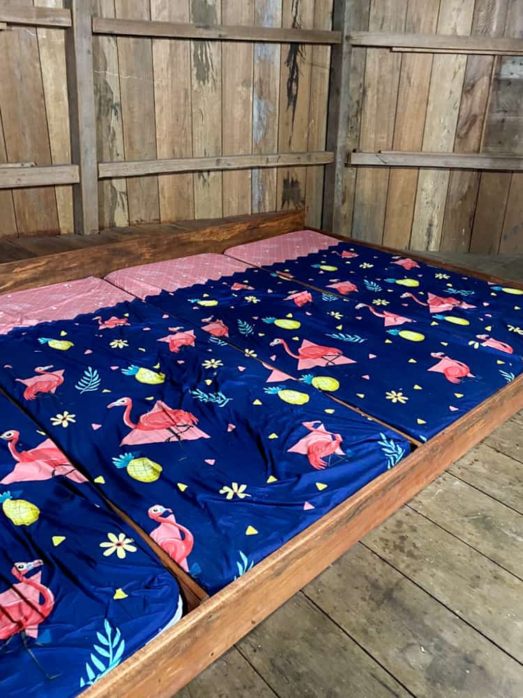 a wooden bed frame on the floor containing four beds with no space in between. The beds have pink sheets and blue blankets with flamingos on them. These are how travelers sleep in guest houses in Wae Rebo traditional village.