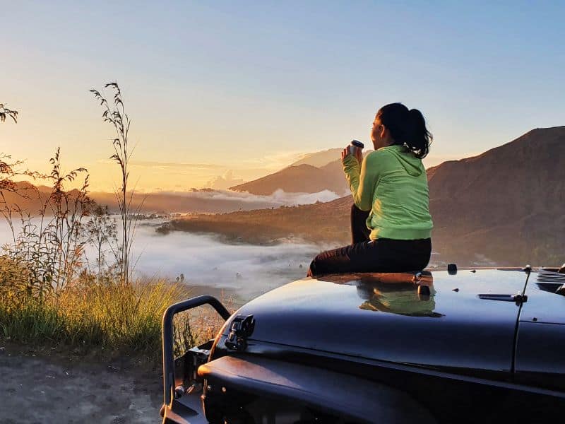A traveler enjoys a cup of coffee while sitting on the hood of a jeep as the sun rises over Mt. Batur volcano in the distance. Taking a jeep up the caldera of Mt. Batur is one of the most popular Bali volcano tours.