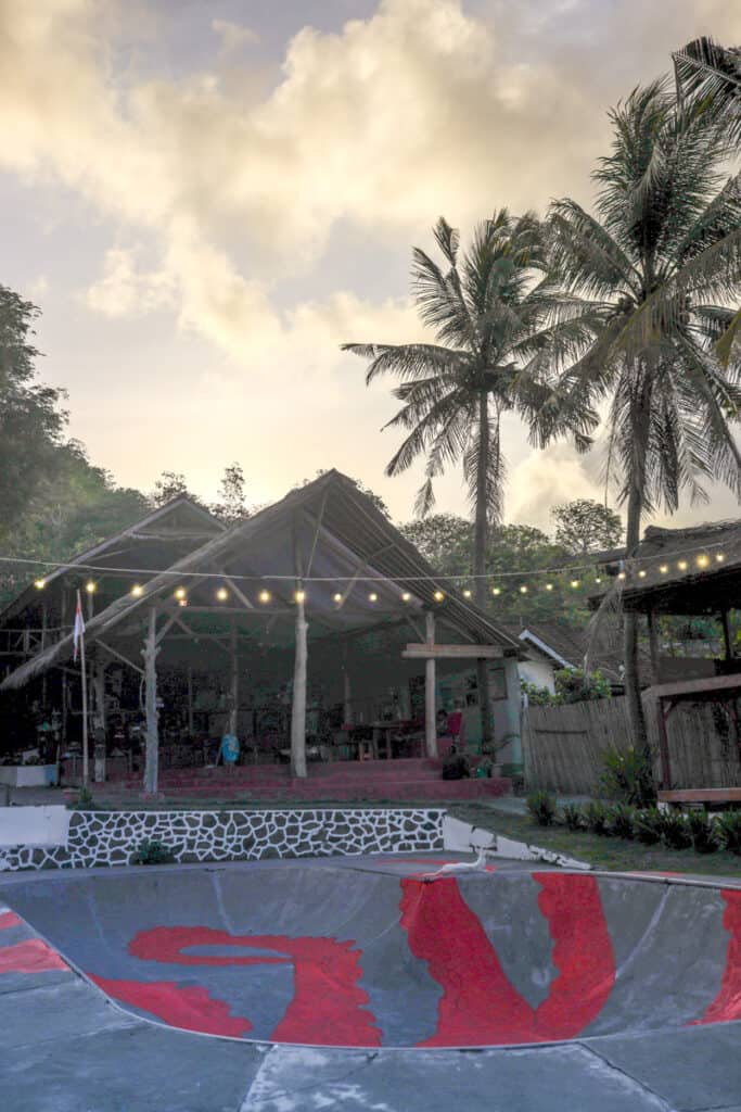 The exterior of a lombok long house at dusk with the sun setting behind it. To the right of the image, you can see palm trees, and in the foreground there is a skate bowl and some twinkle lights. 