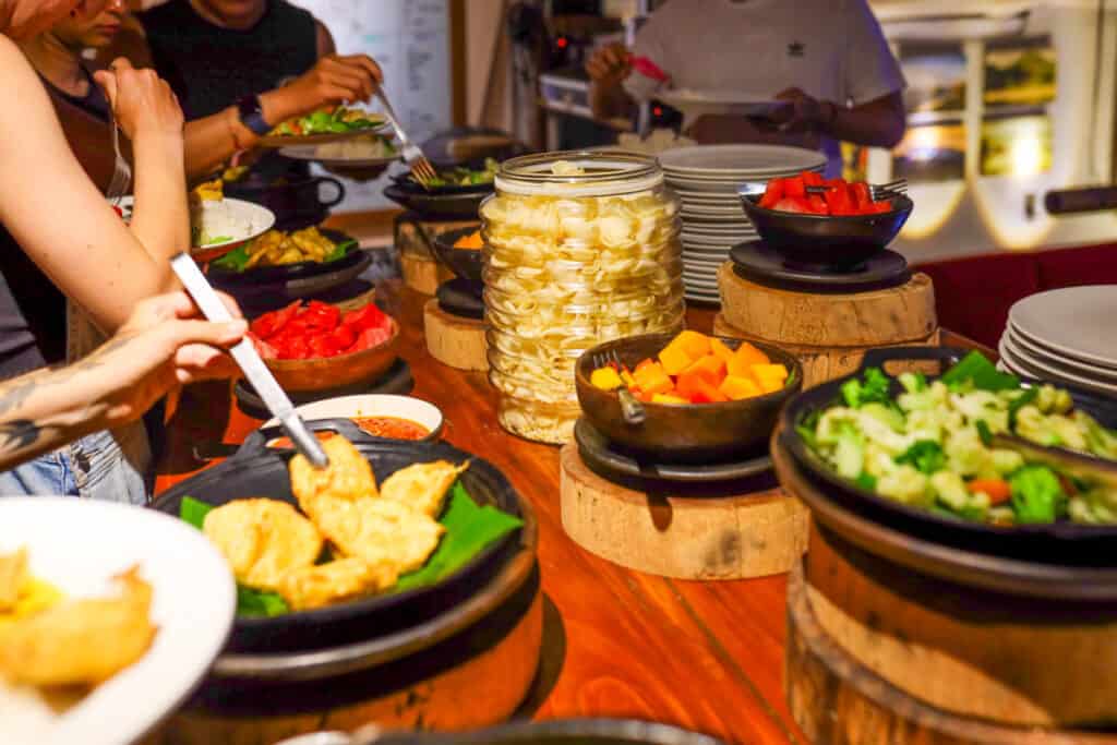 An array of food on a buffet spread out on a wooden table. The food is presented in black stone bowls, with one clear glass container of chips.