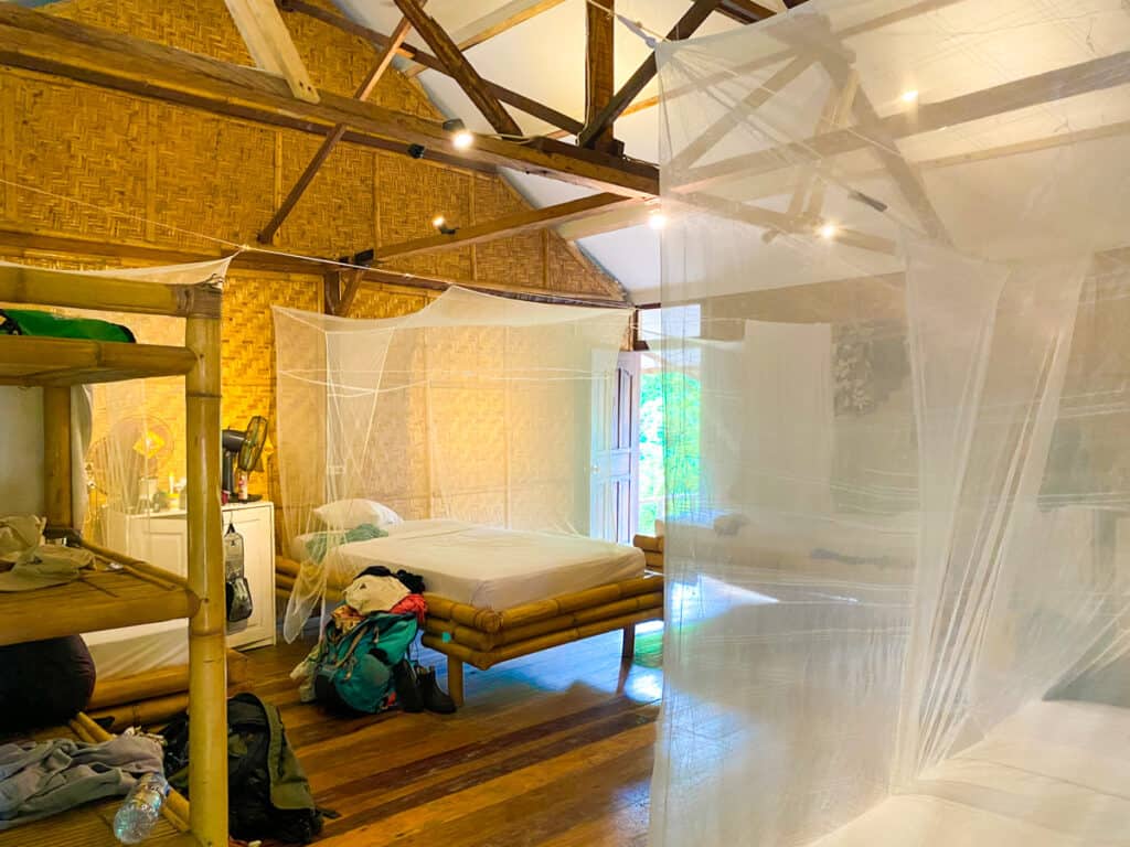 A dormitory room with white mosquito nets around all of the beds. The walls are made of bamboo, with beams on the ceiling also made of bamboo poles.