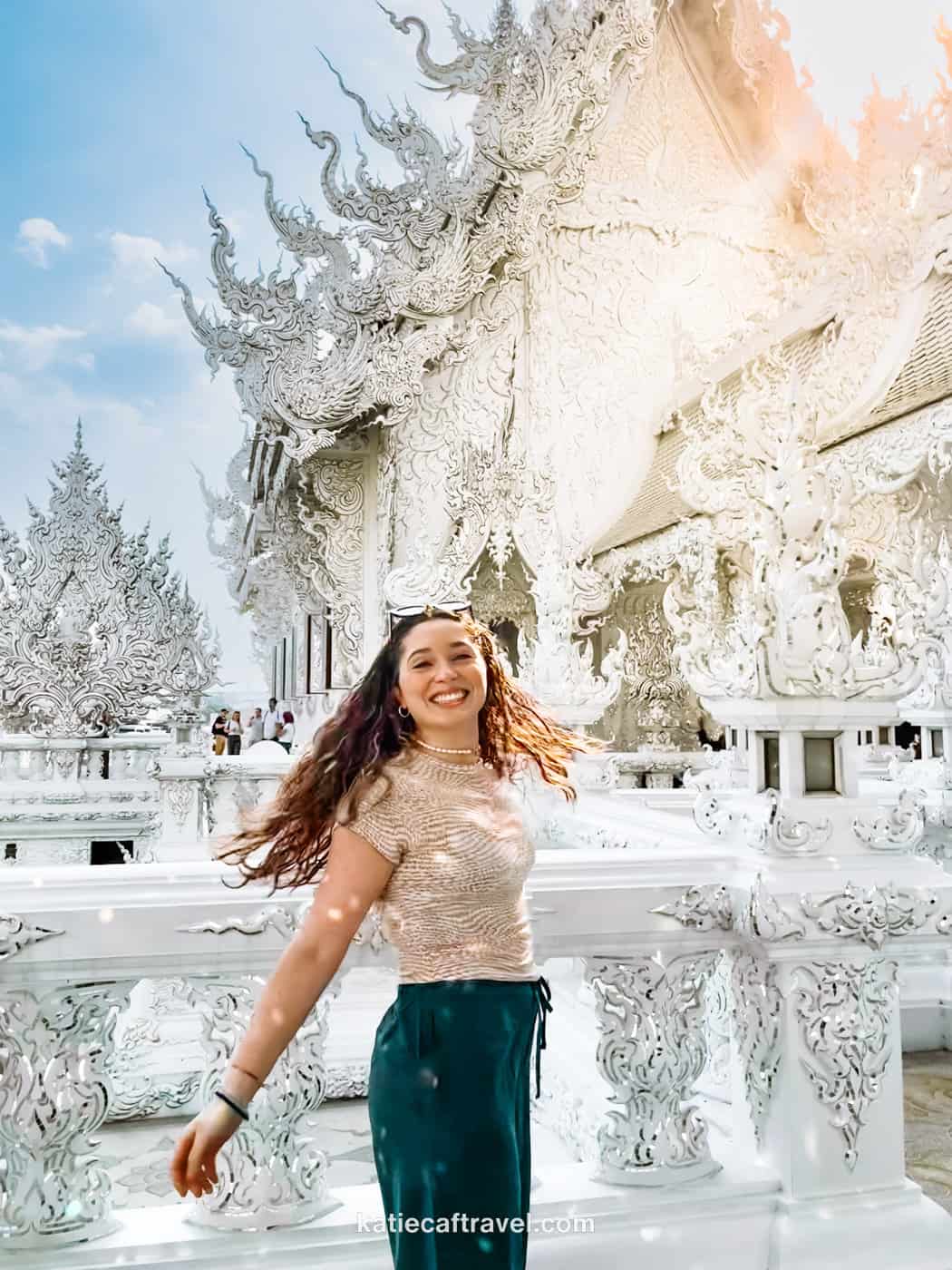 Blogger Katie Caf poses in front of the white temple in Chiang Rai on a sunny day with blue skies.