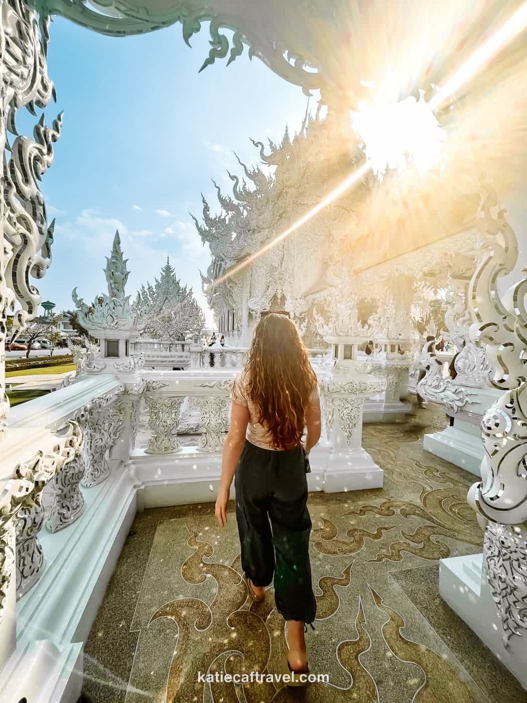 Blogger Katie Caf faces away from the camera walking towards white, intricately carved balcony, in front of the White Temple in Chiang Rai, Thailand.