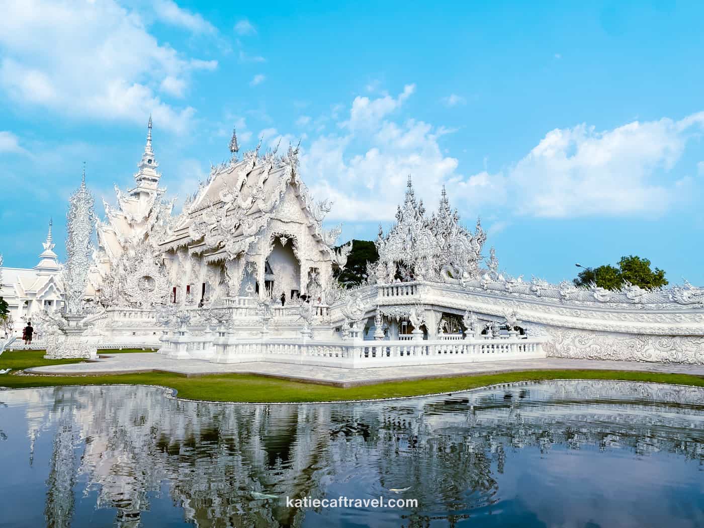 The exterior of the White Temple in Chiang Rai photographed on a sunny day with the reflection in the lake in front of it.